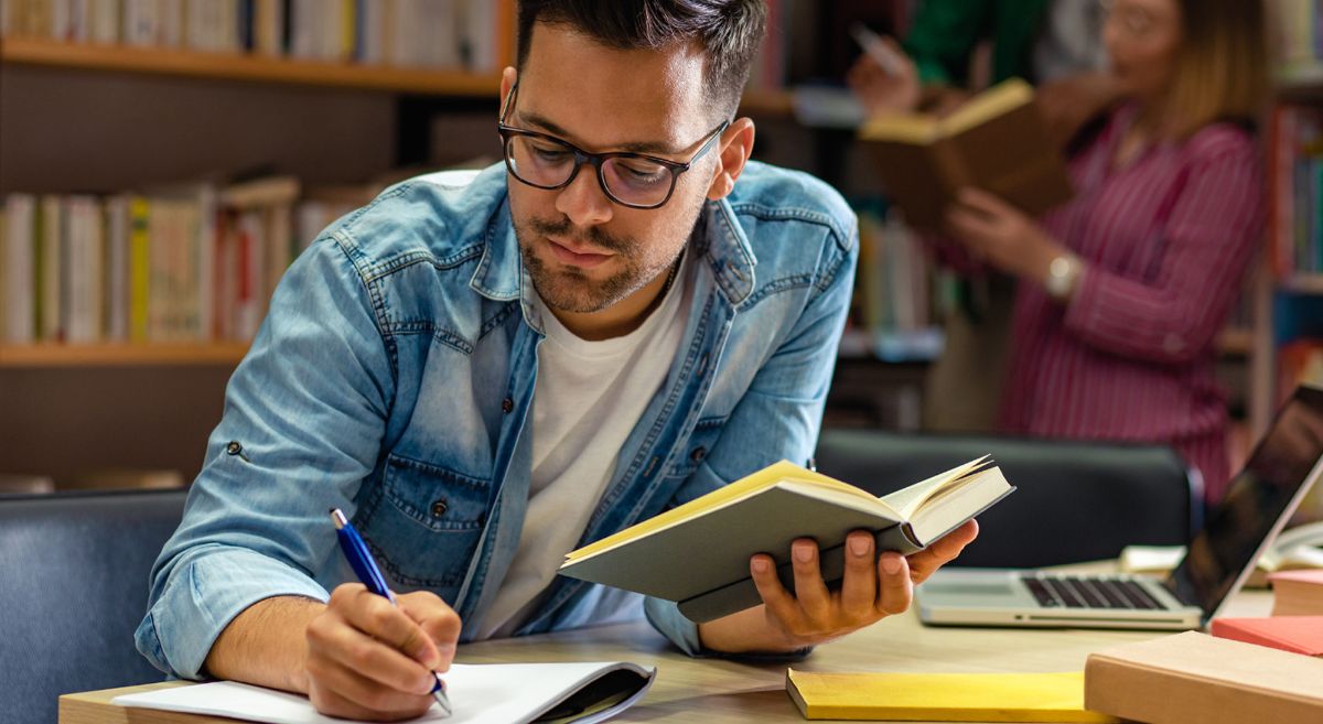 Young male, wearing blue denim shirt and wearing glasses,  holding book and writing on pad in library